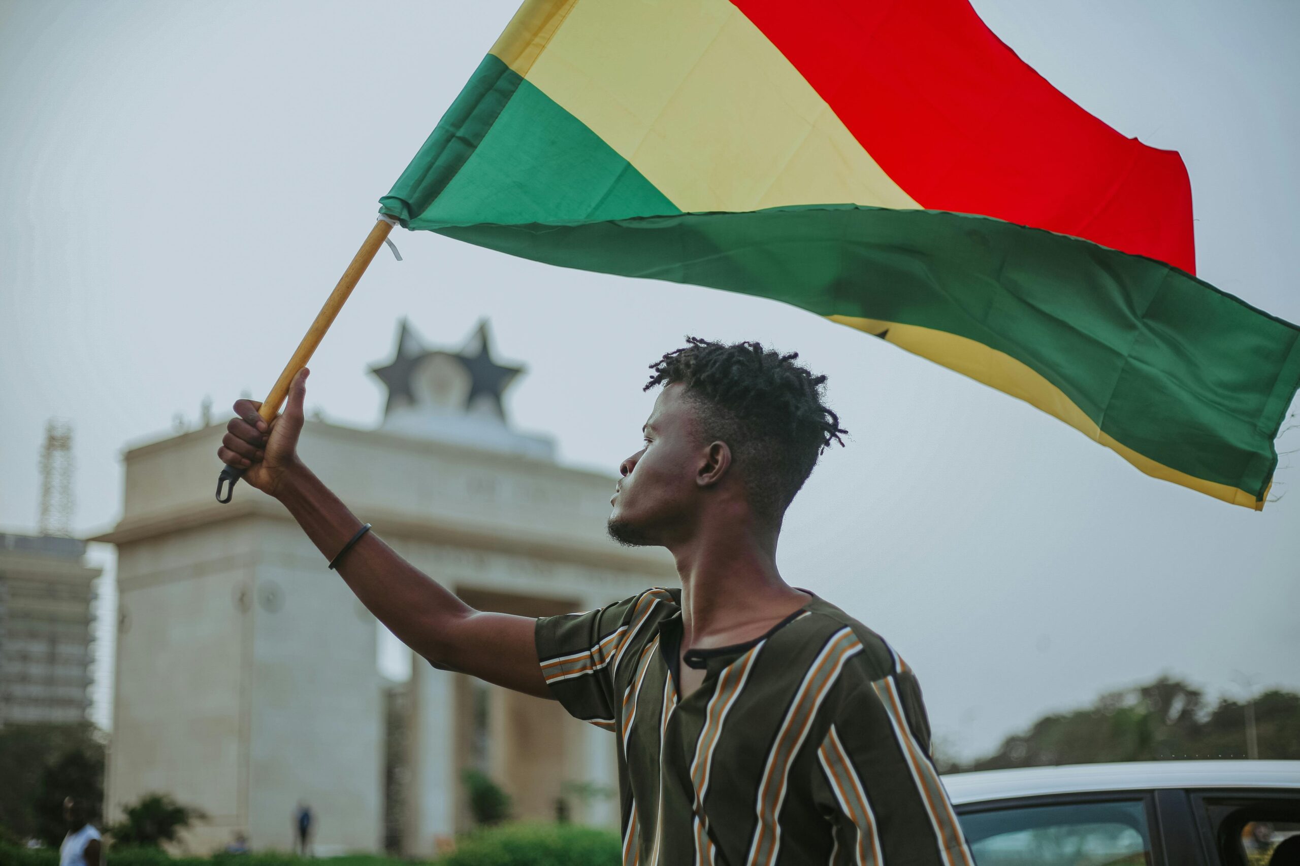 Home African male with dreadlocks raising flag of Ghana country with colorful stripes while looking away in town